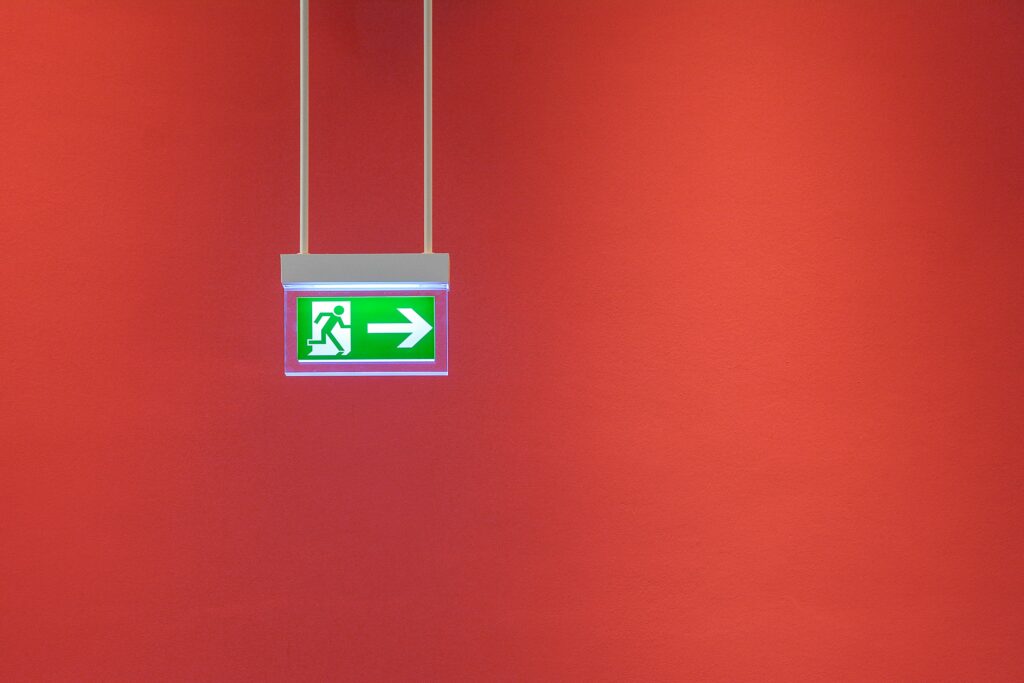 Green emergency exit sign with arrow on vibrant red wall, indicating direction for safety.